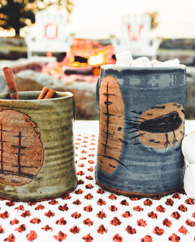 A green mug to the left and a right mug to the right sit in front of a bonfire with chairs in the background that have the letters 'L, O, V, E' printed on them.