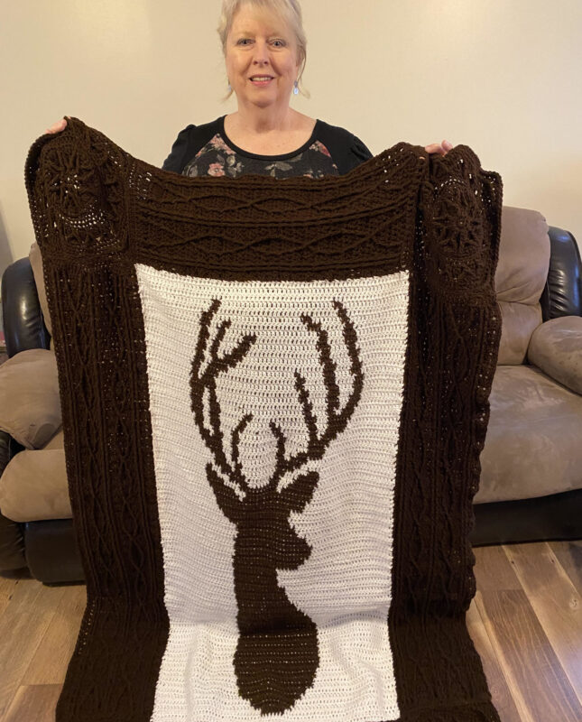 A woman holding a brown blanket with a white square containing a deer head crocheted in the middle.