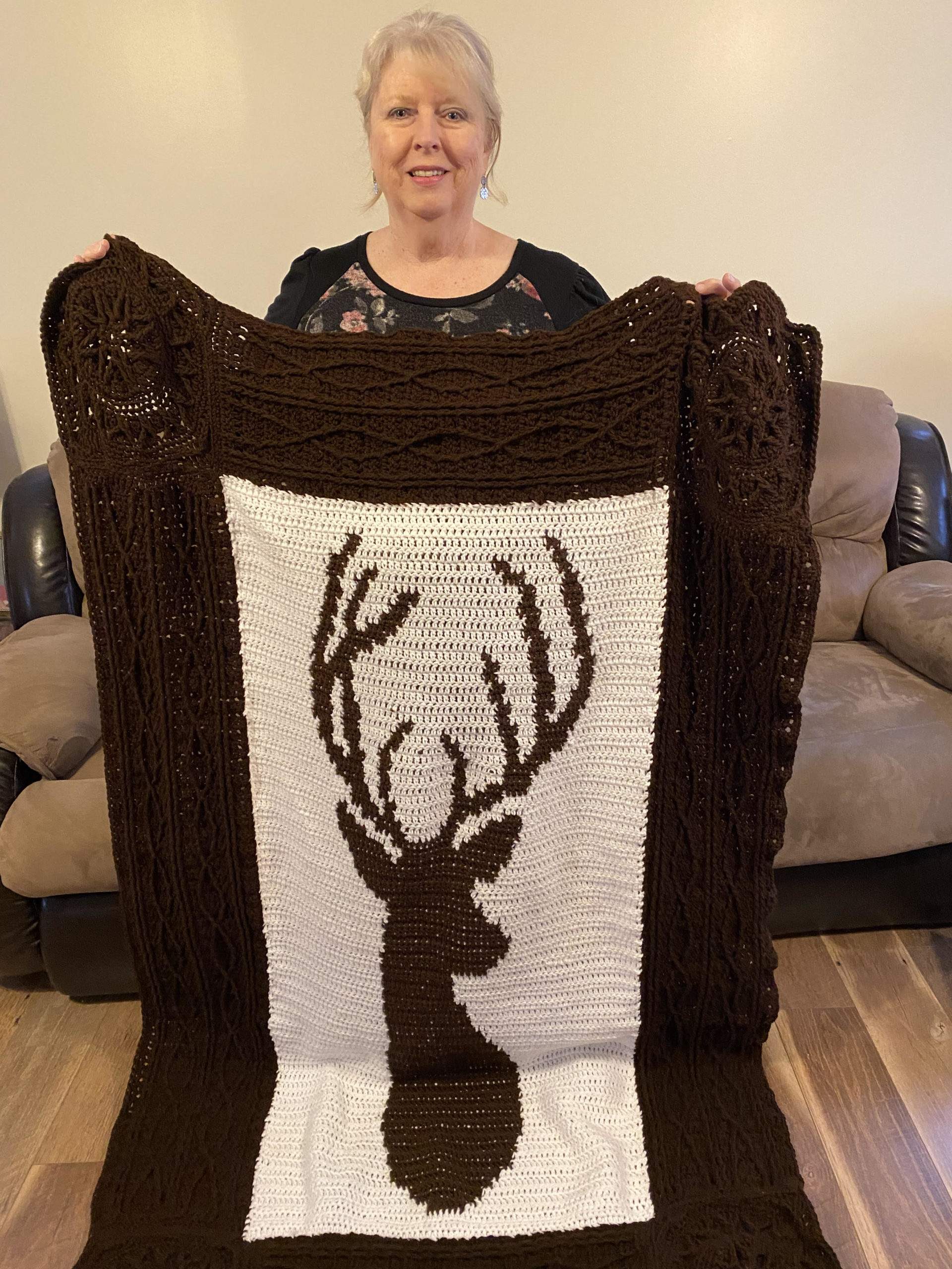 A woman holding a brown blanket with a white square containing a deer head crocheted in the middle.